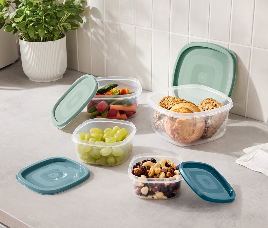 Na lince stojí čtyři průhledné nádoby s modrými víčky, naplněné hrozny, nakrájenou zeleninou, ořechy a sušenkami.
