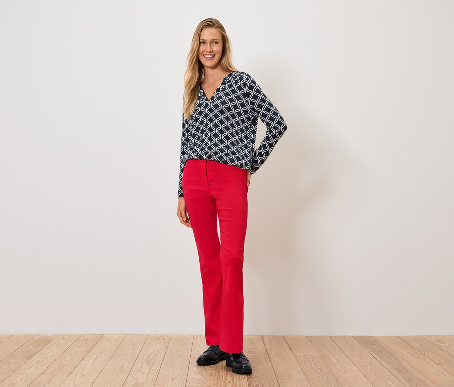 Žena v modro-bílém topu a červených kalhotách stojí na dřevěné podlaze.