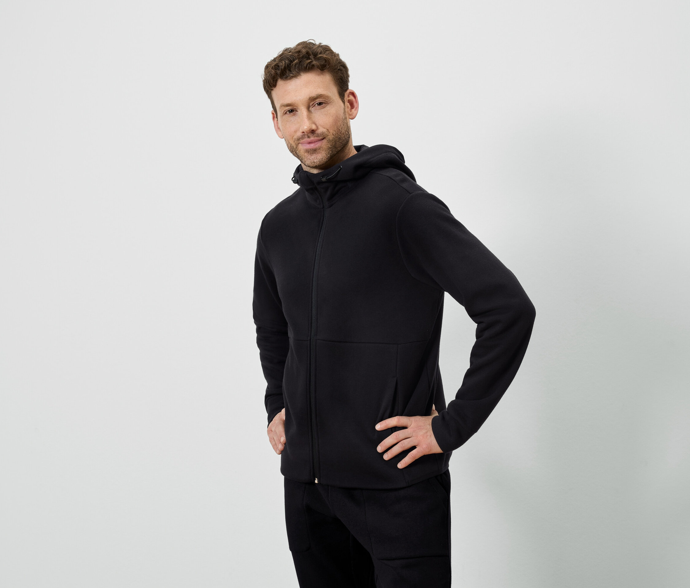 Muž pózuje na bílém pozadí. Má na sobě černou mikinu s kapucí a černé kalhoty.