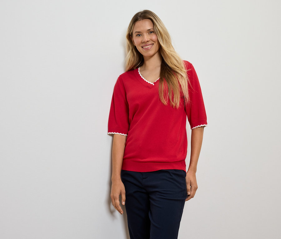 Žena s blond vlasy pózuje v červeném svetru a tmavě modrých kalhotách.