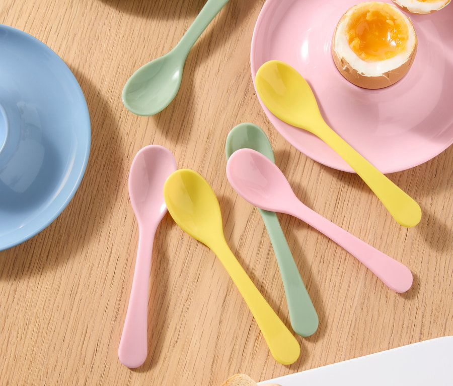 Pohled shora na stůl s vařeným vejcem v kalíšku na vejce, půlkou vařeného vejce na talíři, barevnými lžičkami a toastem.