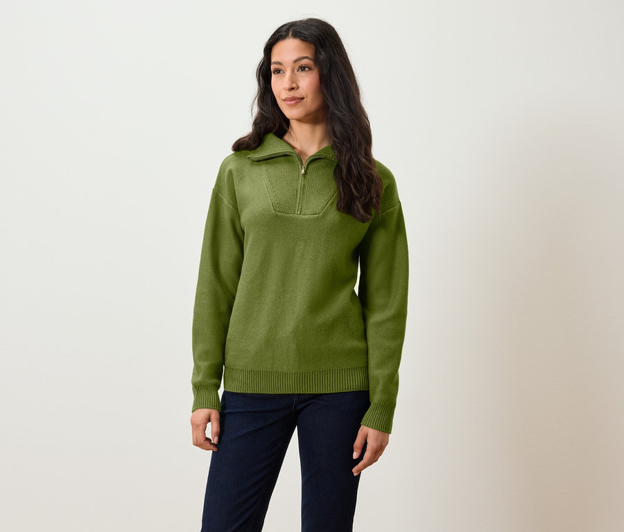 Žena stojí a má na sobě olivově zelený svetr s krátkým zipem a tmavě modré kalhoty.