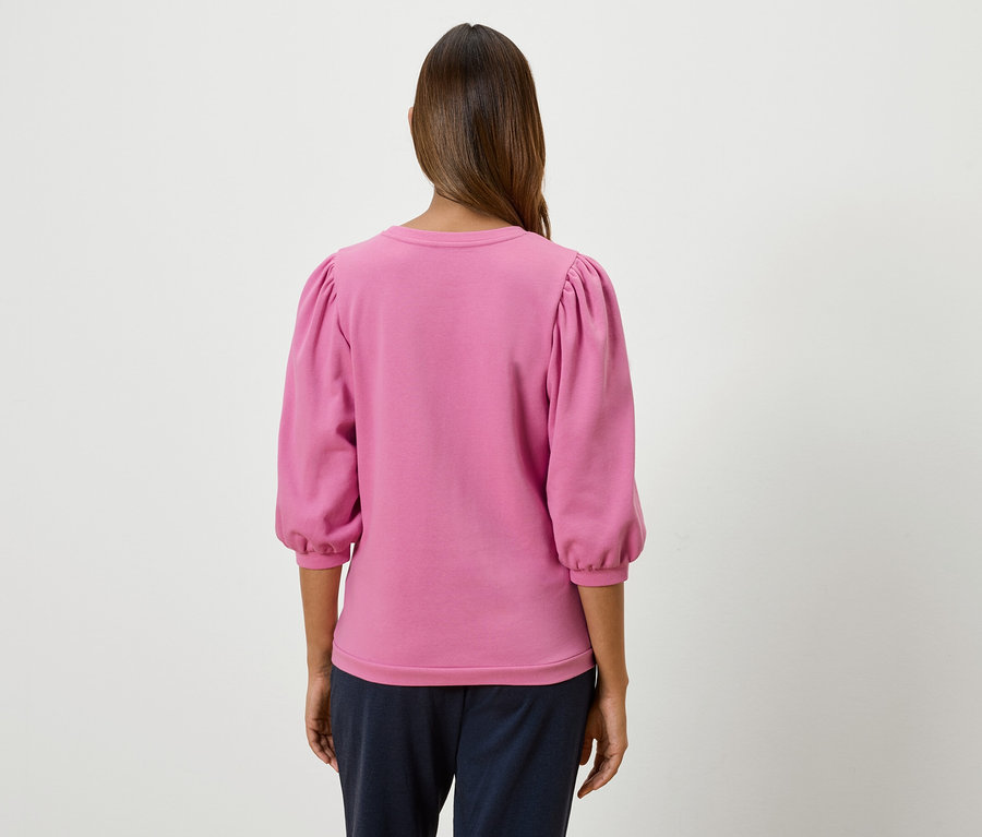Žena stojí zády a má na sobě růžovou mikinu s tříčtvrtečními nabíranými rukávy.