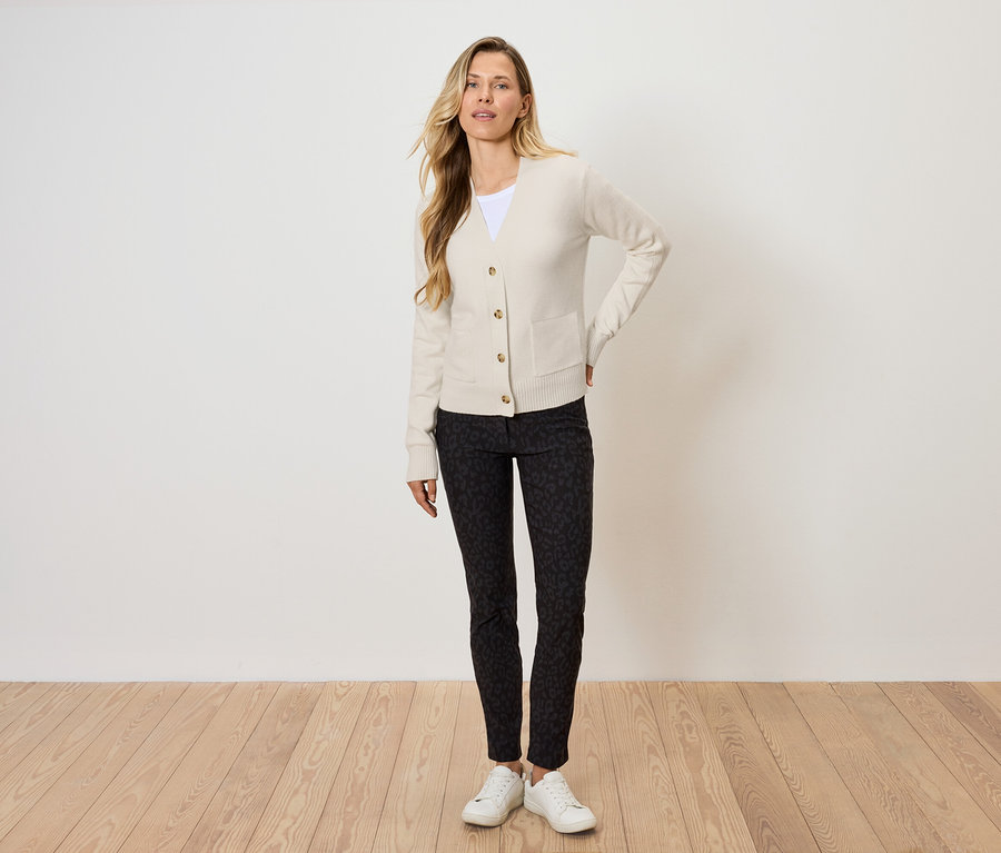 Blonďatá žena pózuje v krémovém kardiganu, černých vzorovaných kalhotách a bílých teniskách.