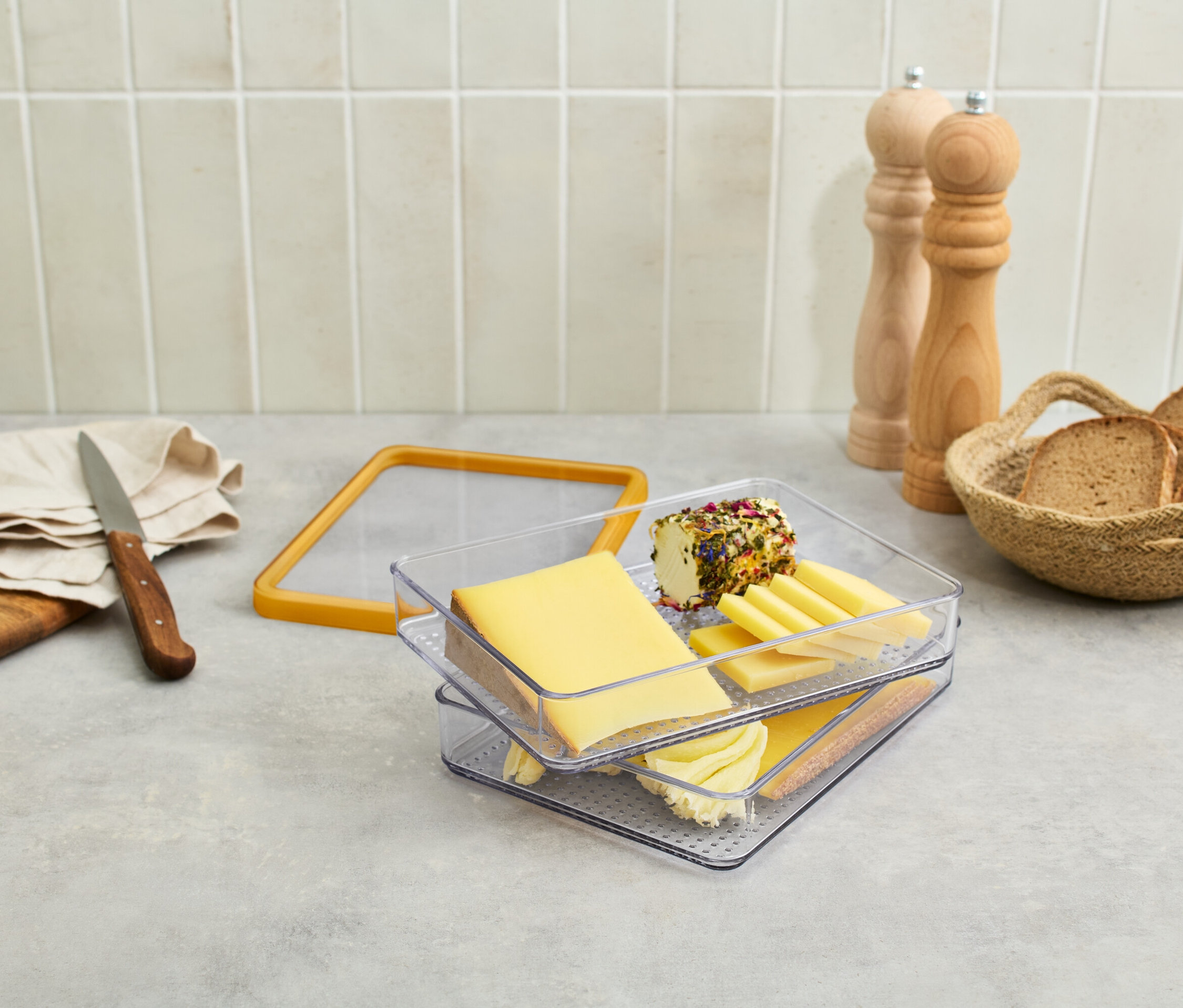 Na stole je sýr v průhledné nádobě, hrozny, nůž, chléb v košíku a mlýnky na koření.