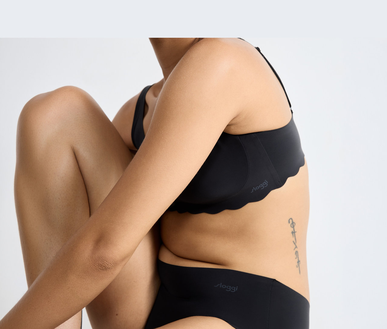 Osoba v černé měkké podprsence sloggi ZERO Microfibre 2.0 Soft bra s vlnitým okrajem a odpovídajícími černými kalhotkami sloggi, sedící se skrčenými koleny a zkříženýma rukama. Na boku je vidět tetování.