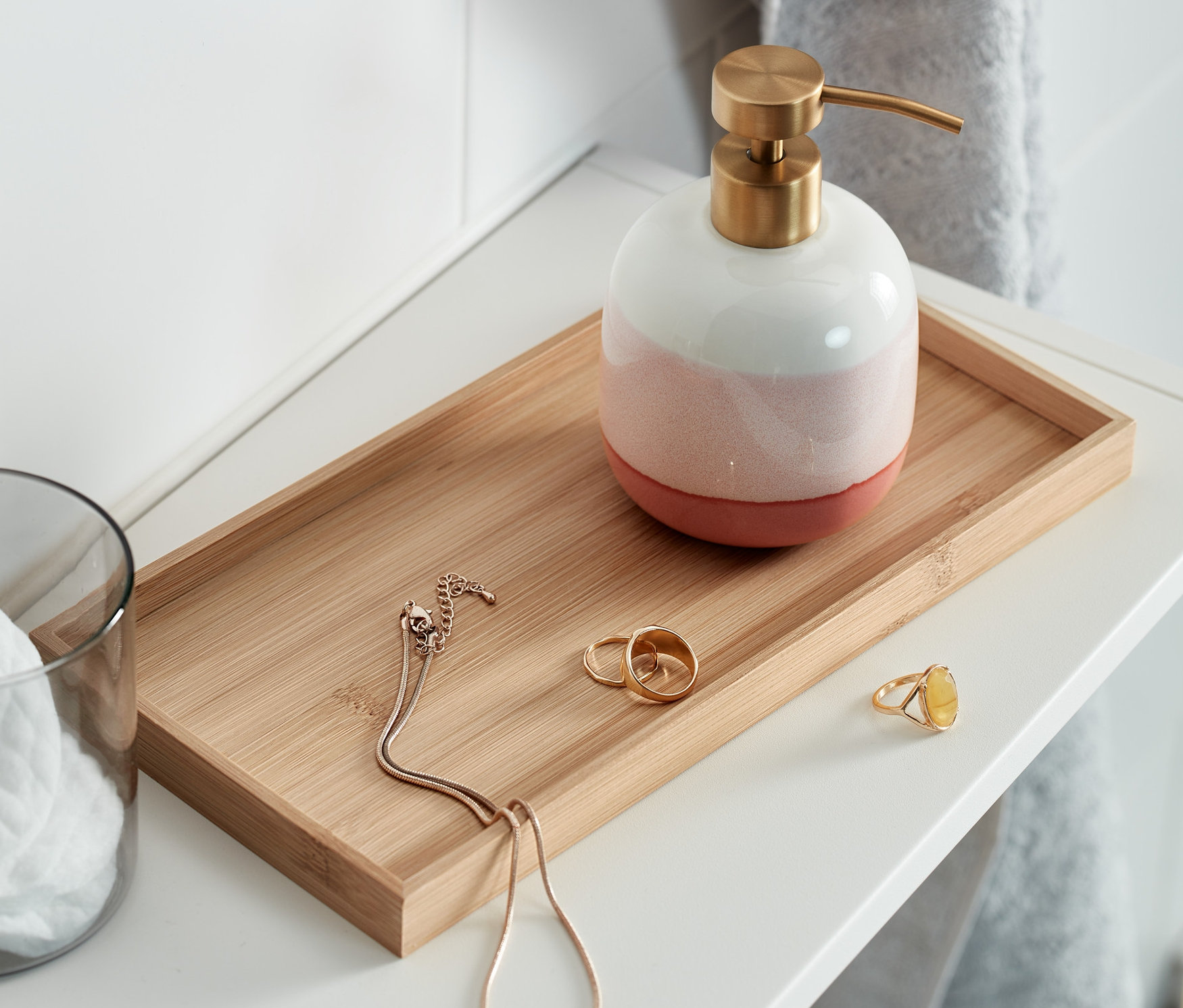 Na bílé polici je ozdobný podnos s dávkovačem mýdla, sklenicí s vatovými tampony a šperky.