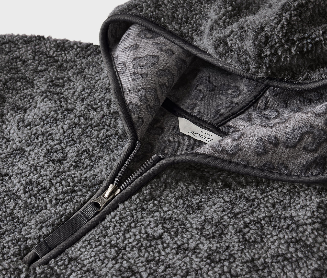 Detail šedé chlupaté mikiny s kapucí se zipem a šedým zvířecím vzorem na vnitřní straně kapuce.