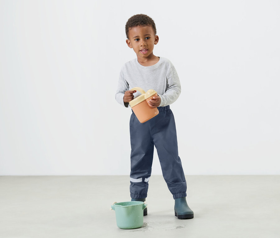 Malý chlapec drží v rukou oranžový kbelík a má na sobě modré nepromokavé kalhoty a holínky.