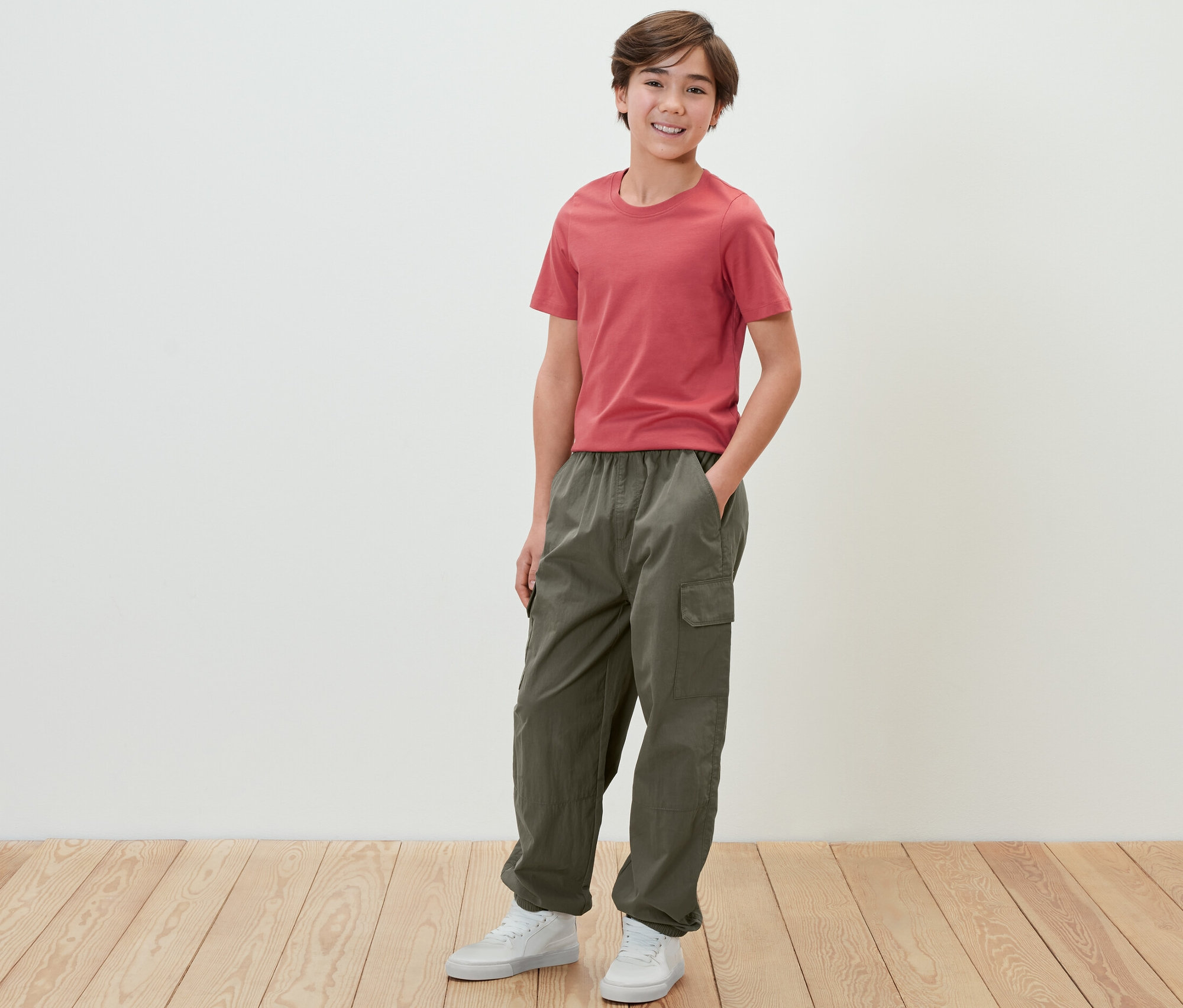 Chlapec v červeném tričku a dětských cargo kalhotách stojí před bílou zdí.