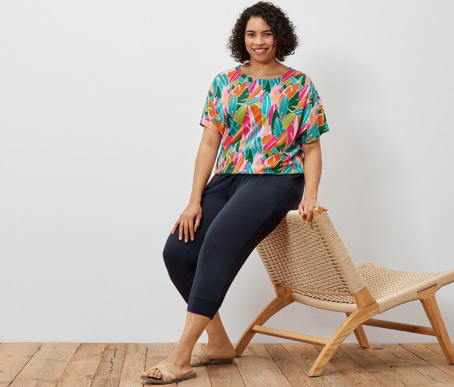 Žena sedí na proutěném křesle a má na sobě tmavě modré 3/4 žerzejové kalhoty a halenkové tričko se spadlými rameny s celoplošným potiskem.