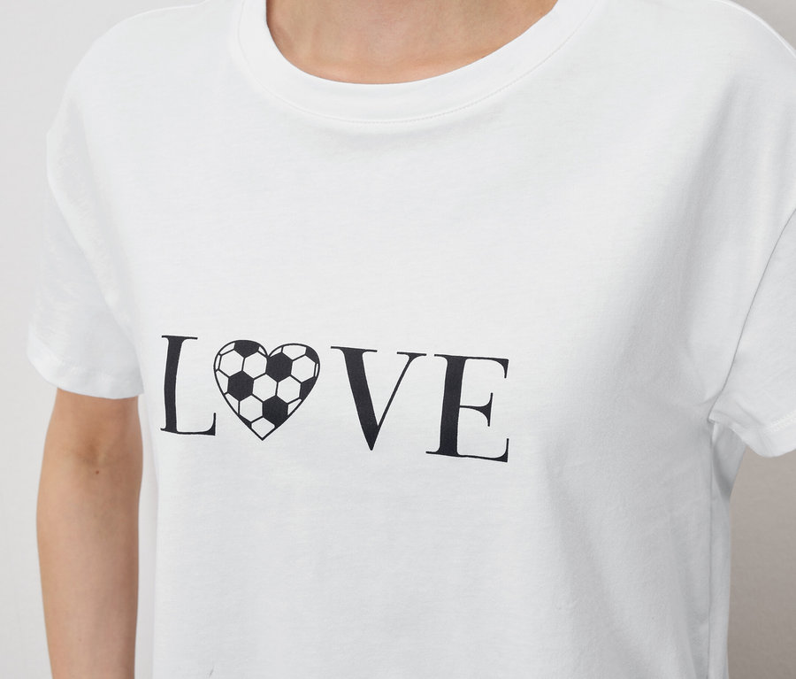 Detailní záběr na osobu v bílém tričku s nápisem „LOVE“, kde je písmeno „O“ nahrazeno srdcem ve tvaru fotbalového míče.