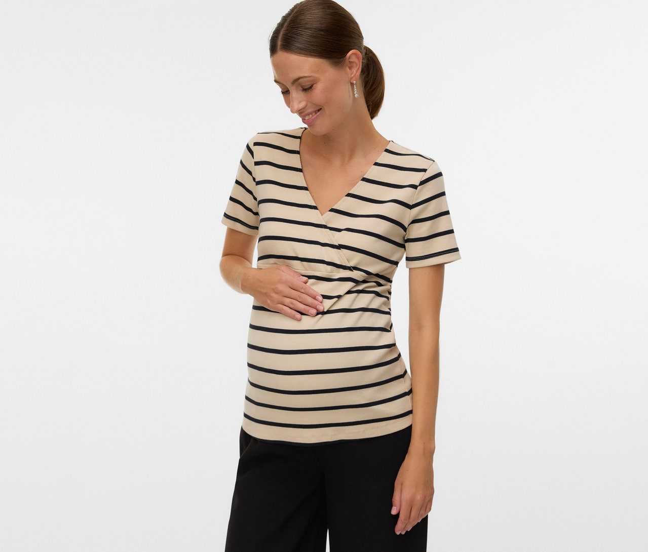 Žena s culíkem má na sobě těhotenské tričko MAMALICIOUS »MLLANE TESS« s proužky a černé kalhoty a dotýká se břicha.