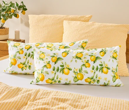 Na posteli leží dva polštáře se vzorem citronů před dvěma žlutými polštáři. Vlevo stojí citronová rostlina vedle svíčky.