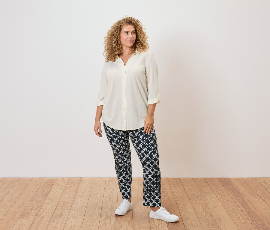 Žena s kudrnatými vlasy má na sobě krémovou blůzu, vzorované kalhoty a bílé tenisky.