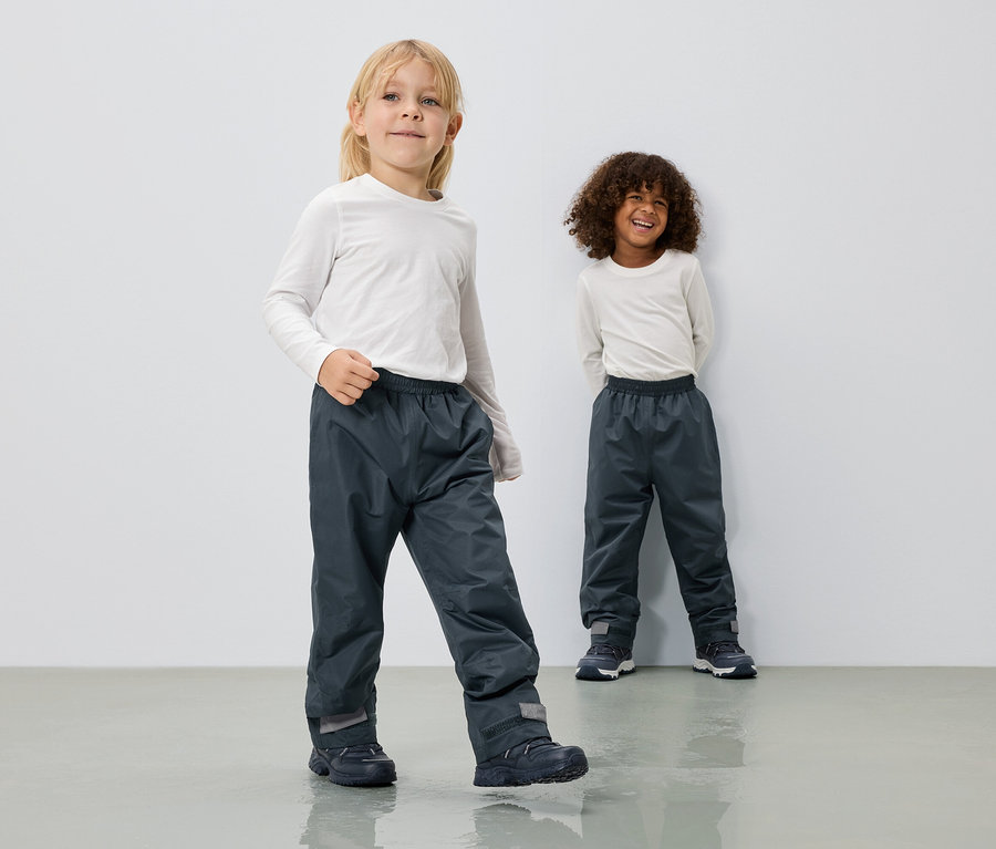 Dvě děti mají na sobě dlouhé šedé nepromokavé kalhoty a bílé trička s dlouhým rukávem před bílou stěnou.