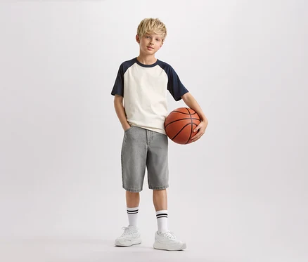 Chlapec s blond vlasy drží basketbalový míč, má na sobě světlé tričko s tmavě modrými rukávy, šedé šortky a bílé ponožky s černými pruhy.