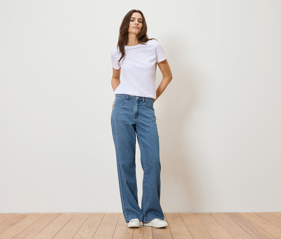 Žena stojí před bílou zdí a má na sobě bílé tričko a modré džíny.