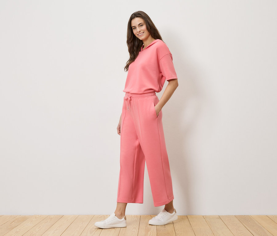 Žena má na sobě korálovou teplákovou soupravu s mikinou s kapucí a culottes a bílé tenisky.