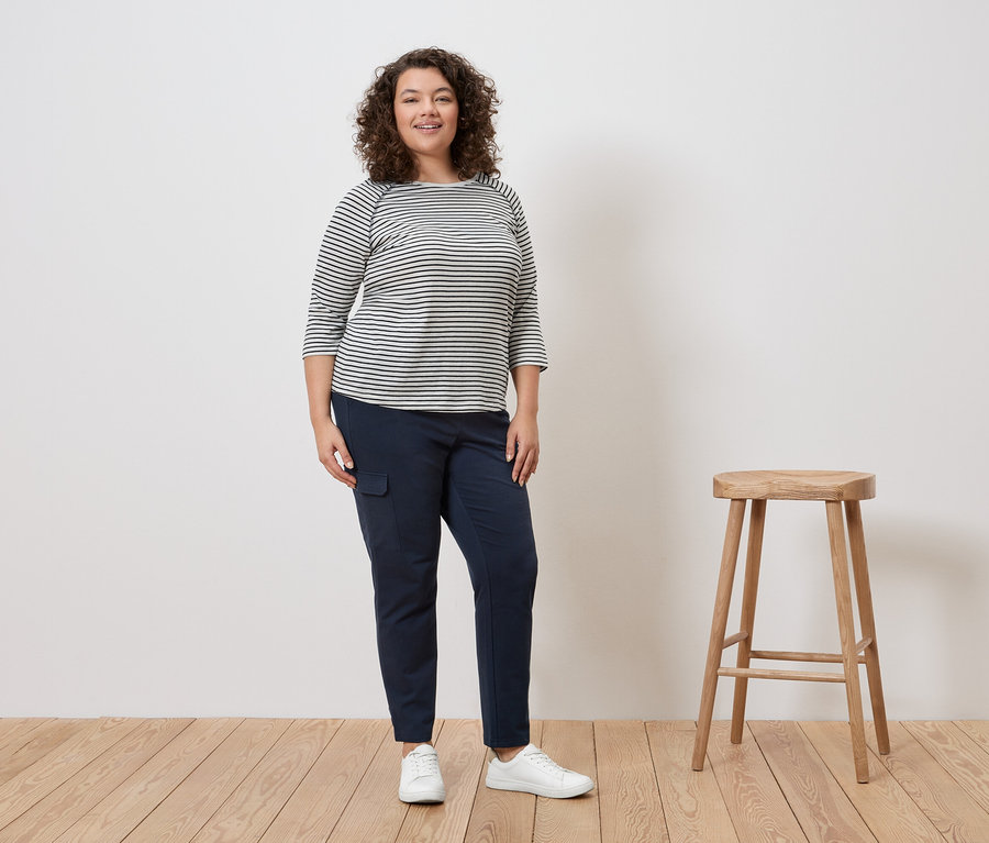 Žena s kudrnatými vlasy stojí v pruhovaném topu, modrých kalhotách a bílých teniskách vedle stoličky.