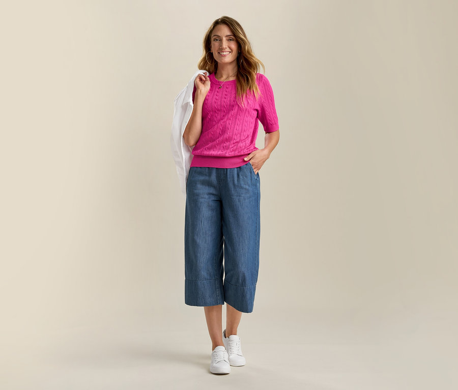 Usmívající se žena má na sobě džínovou bundu a růžový svetr z jemného úpletu s copánkovým vzorem. Má na sobě modré culottes a bílé tenisky.