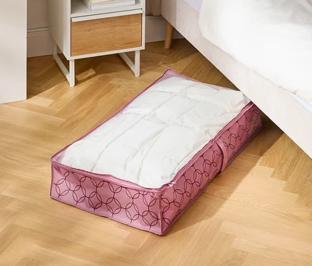Na dřevěné podlaze leží růžová úložná taška pod postelí vedle nočního stolku.