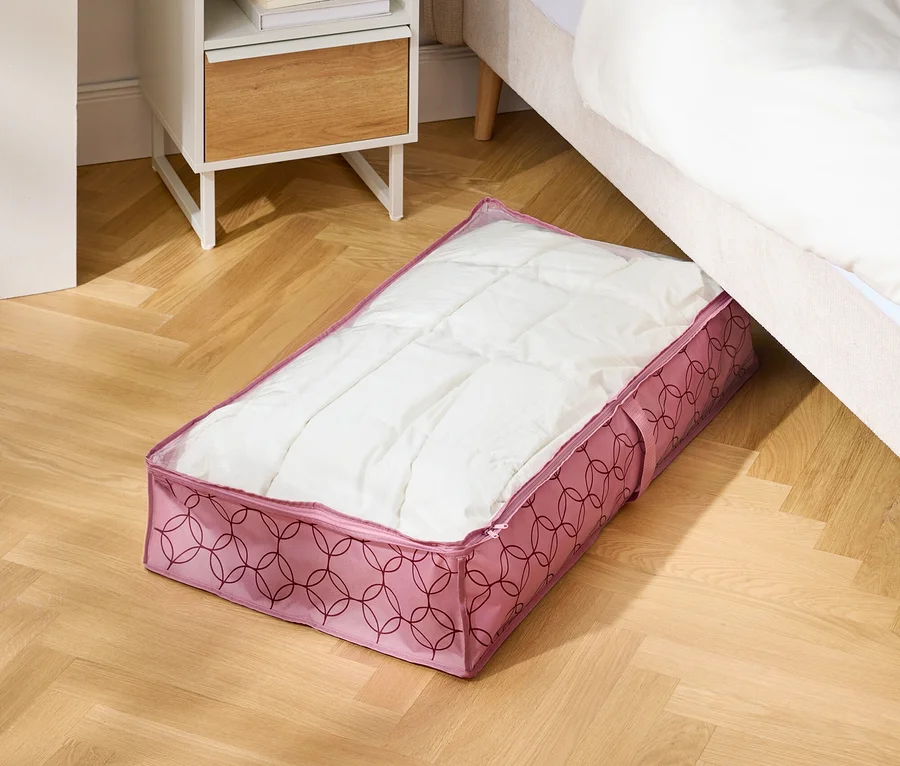 Na dřevěné podlaze leží růžová úložná taška pod postelí vedle nočního stolku.
