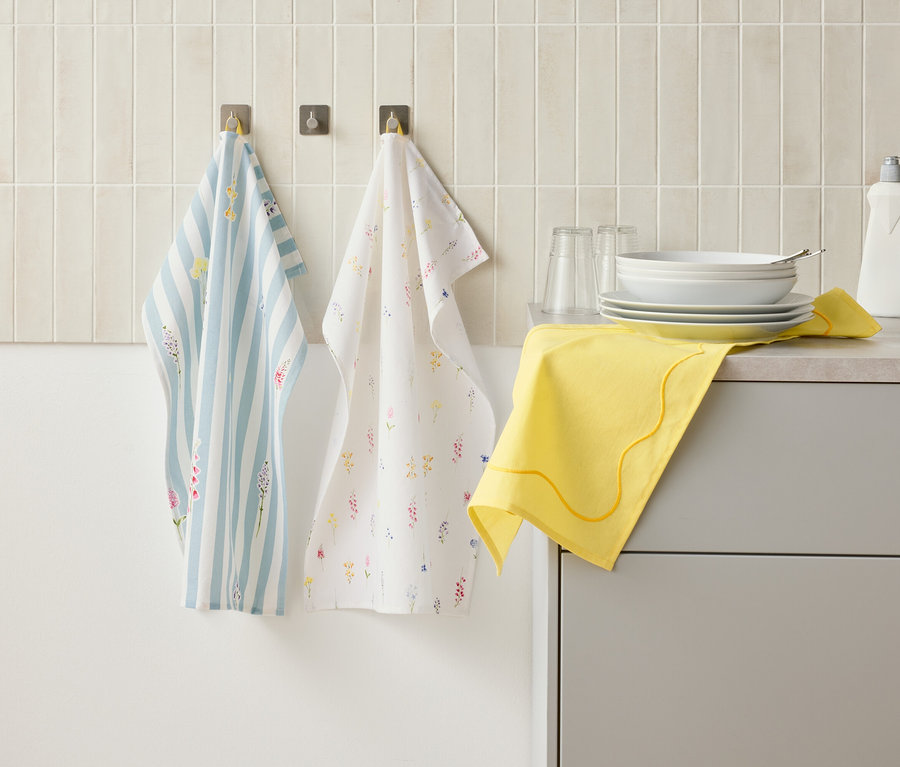 Dvě kuchyňské utěrky visí na háčcích na zdi nad kuchyňskou linkou, na které je nádobí, sklenice, láhev saponátu a žlutý hadřík.