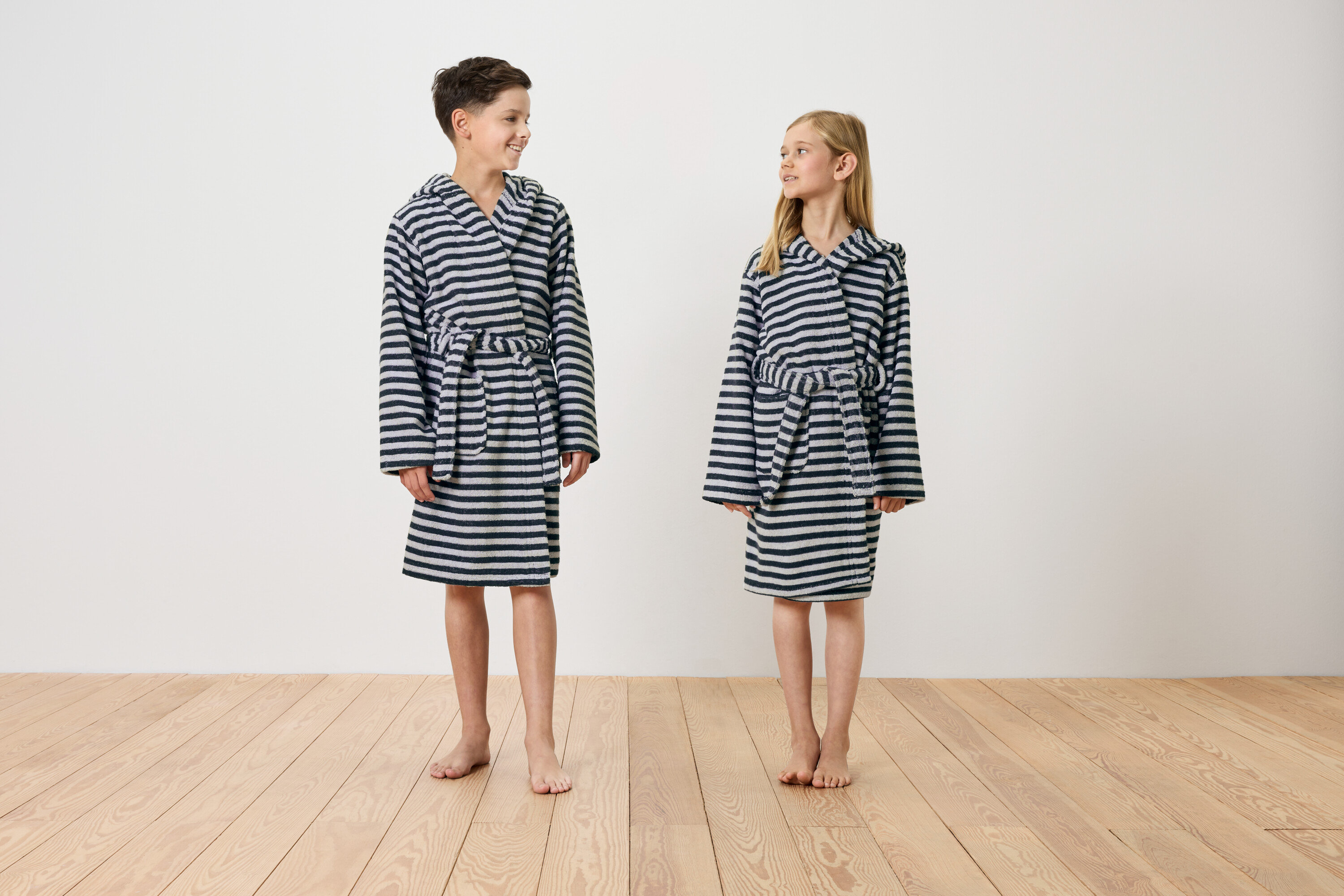 Chlapec a dívka stojí bosí na dřevěné podlaze a mají na sobě pruhované župany s kapucí.