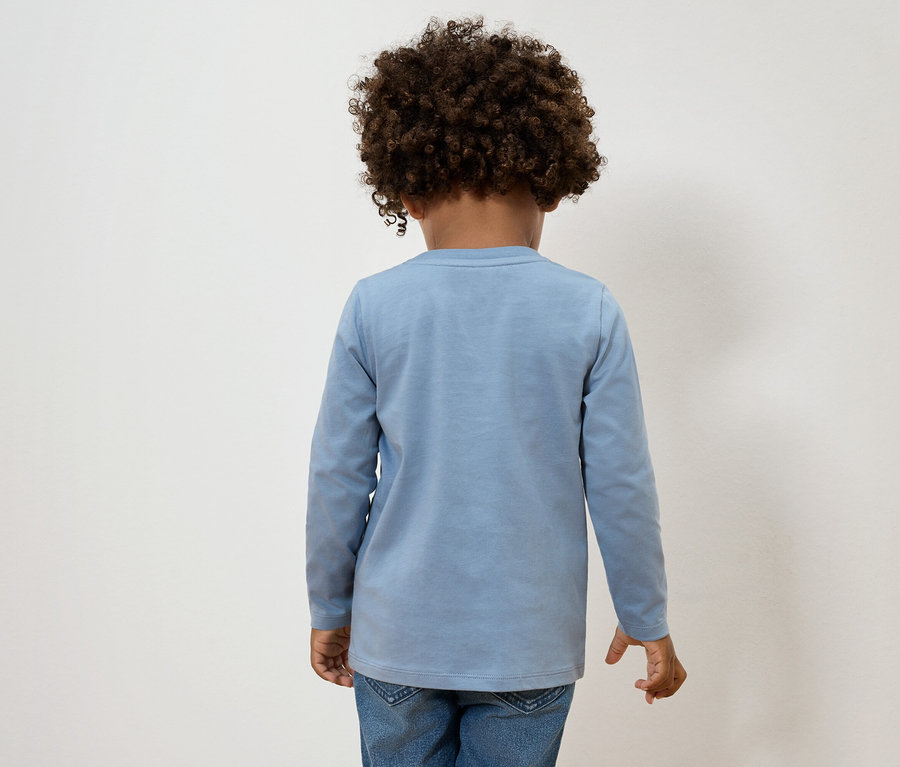 Pohled zezadu na dítě s kudrnatými vlasy, oblečené ve světle modrém tričku s dlouhým rukávem a džíny.