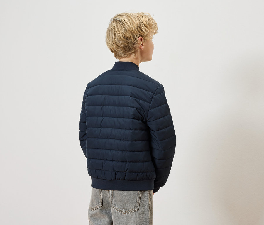 Pohled zezadu na blonďaté dítě v tmavě modré prošívané bundě a šedých džínách na bílém pozadí.