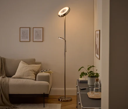 Moderní stříbrná stojací lampa s hlavním světlem směřujícím nahoru a nastavitelnou lampičkou na čtení osvětluje útulný obývací pokoj. Viditelná je béžová pohovka s dekou a polštářem, dva zarámované obrazy s linkovou kresbou a tmavá skříňka s rostlinou a lahví vody.