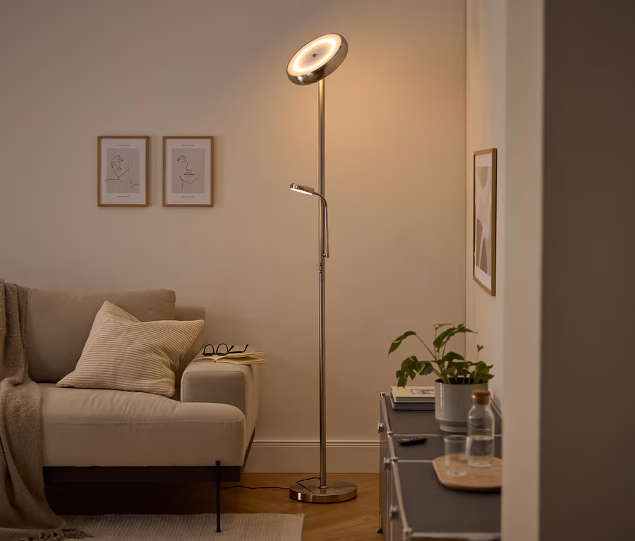 Moderní stříbrná stojací lampa s hlavním světlem směřujícím nahoru a nastavitelnou lampičkou na čtení osvětluje útulný obývací pokoj. Viditelná je béžová pohovka s dekou a polštářem, dva zarámované obrazy s linkovou kresbou a tmavá skříňka s rostlinou a lahví vody.