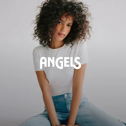 Žena s kudrnatými vlasy v bílém tričku a světle modrých džínách sedí a dívá se do kamery. Na obrázku je překrytý nápis ANGELS.