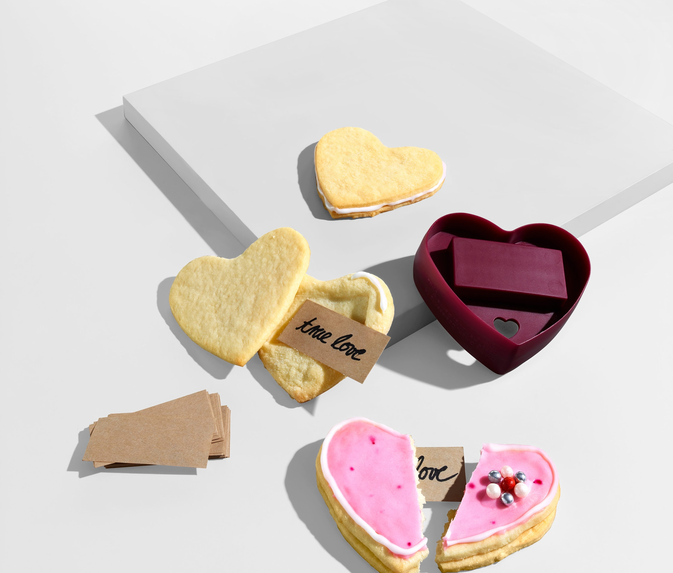 Srdíčkové sušenky a vykrajovátko na sušenky štěstí ve tvaru srdce leží na bílém pozadí.