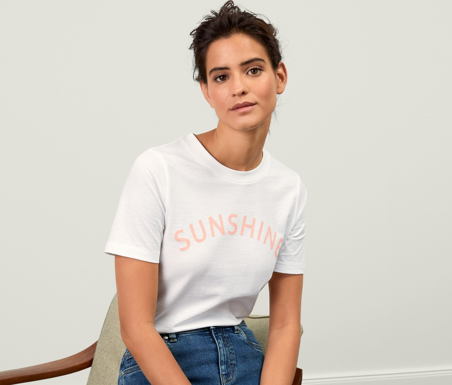 Žena sedí na židli a má na sobě bílé tričko s potiskem.