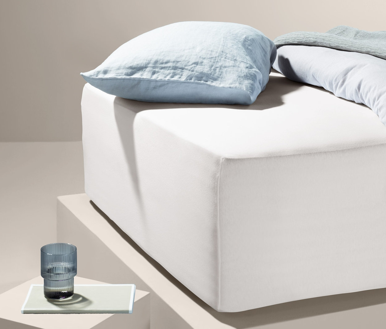 Světle modrý polštář a přikrývka leží na bílém žerzejovém napínacím prostěradle na vysoké matraci.