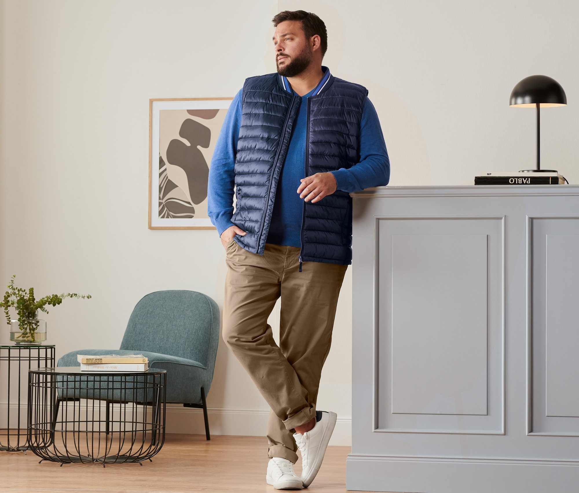 Muž opřený o skříňku má na sobě modrou prošívanou vestu, královsky modrý lněný svetr, kalhoty chino barvy velbloudí srsti a boty sneaker.