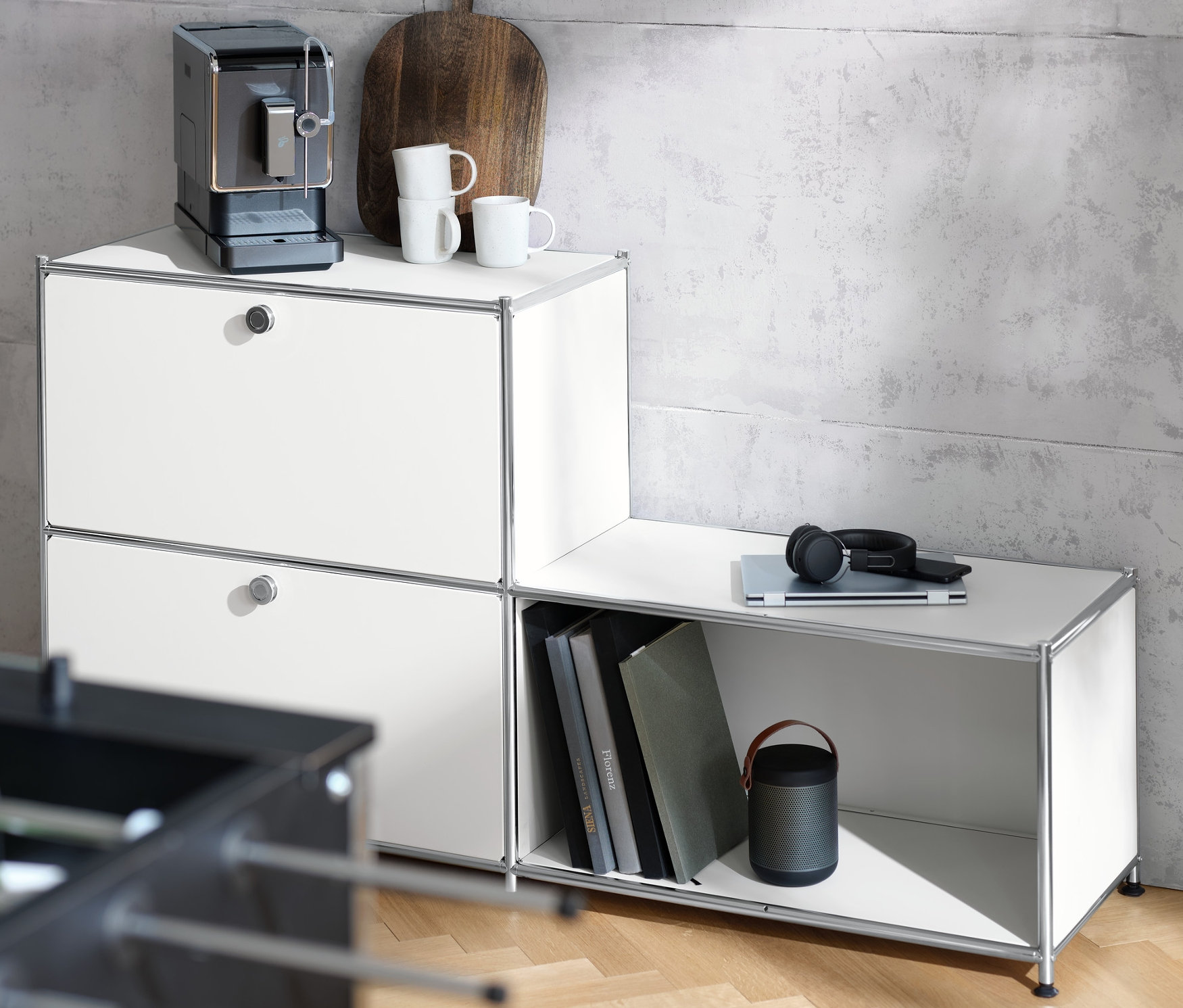 Kávovar stojí na bílém kovovém regálu CN3 se 3 přihrádkami. Na regálu leží také šálek, prkénko a sluchátka.