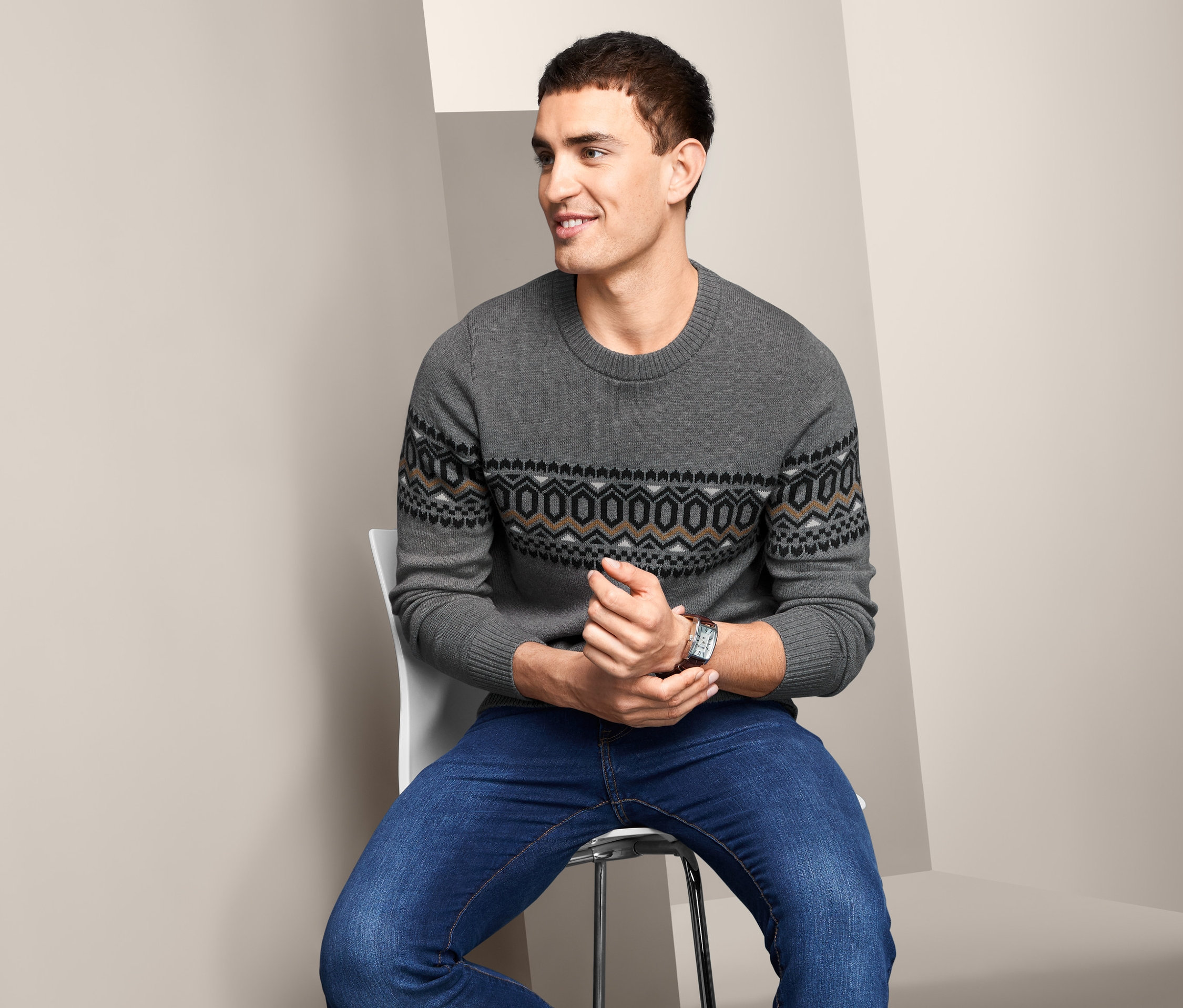 Muž sedí na židli a má na sobě šedý svetr s kulatým výstřihem a džíny.