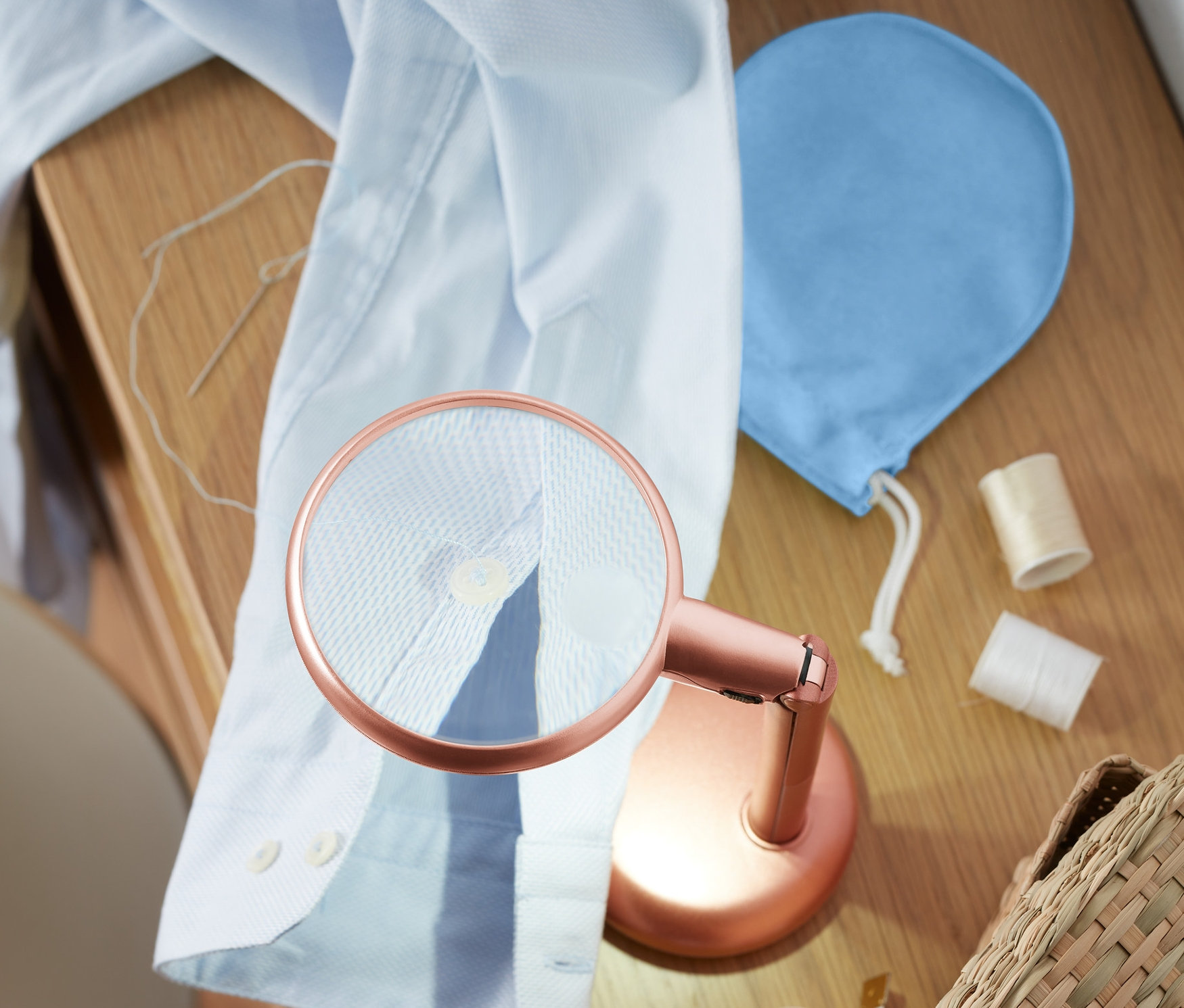 Modrá košile a LED lupa na čtení leží na dřevěném stole vedle šicích potřeb.