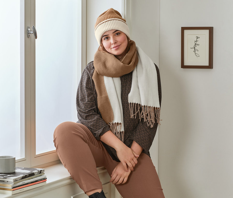 Žena sedí na parapetu v černém halenovém triku s potiskem, koňakových strečových kalhotách, kožených botách, krémovo-koňakové pletené čepici a koňakovo-krémové šále s třásněmi.