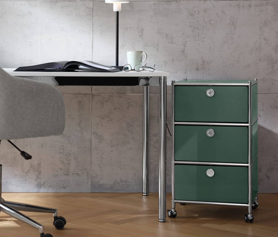 Béžová kancelářská židle stojí u bílého psacího stolu »CN3«, na kterém leží otevřená kniha, kovová stolní lampa, brýle a hrnek. Vedle stolu stojí olivově zelený kontejner na kolečkách CN3 se 3 zásuvkami.