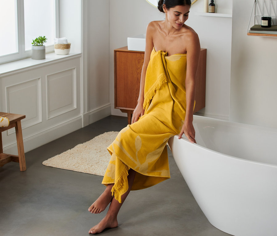 Žena sedí na okraji vany a je zabalená do žluté osušky hammam XL.