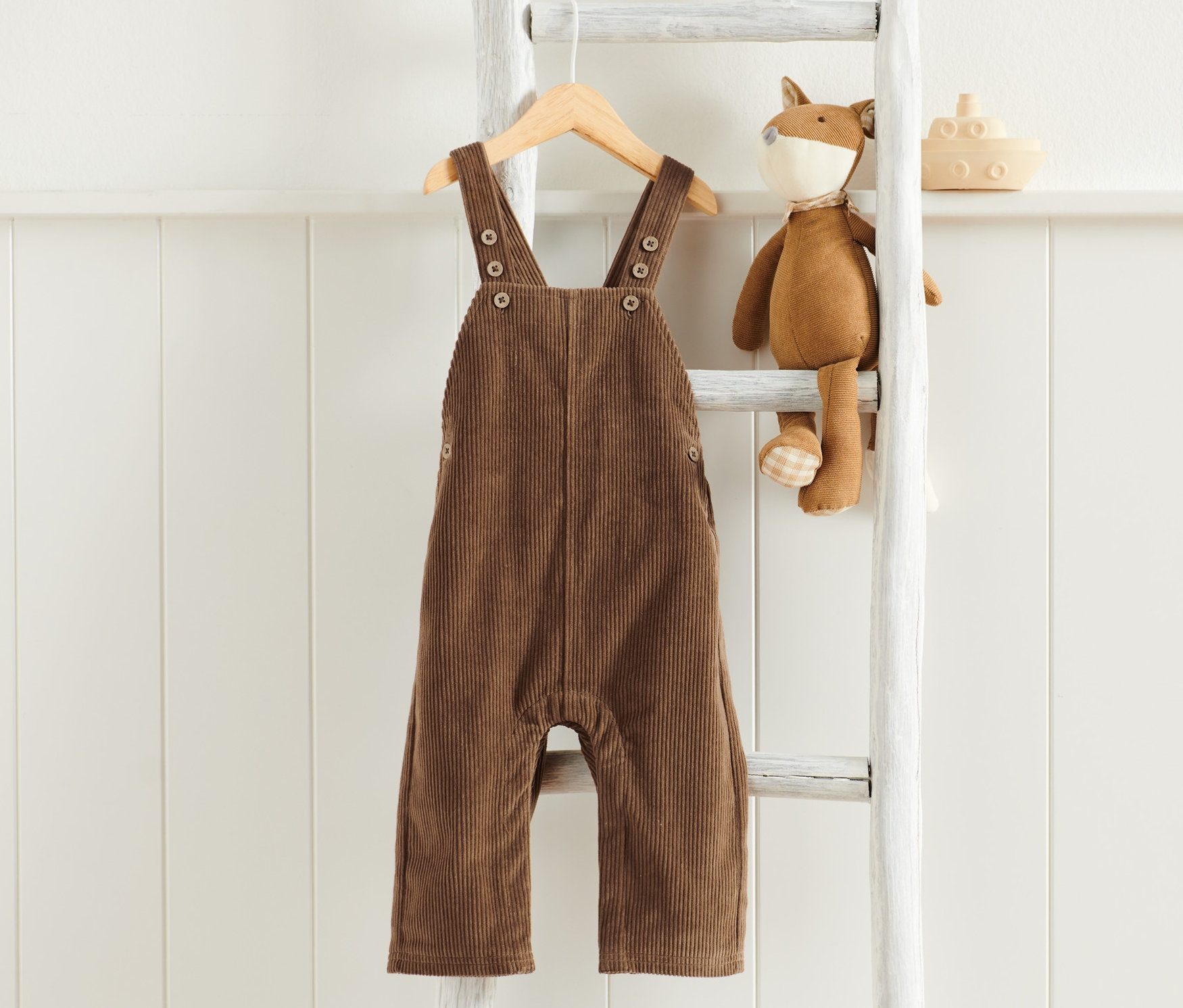 Hnědé manšestrové kalhoty s laclem visí na dřevěném ramínku na bílém žebříku vedle plyšové hračky lišky a dřevěné hračky lodi.