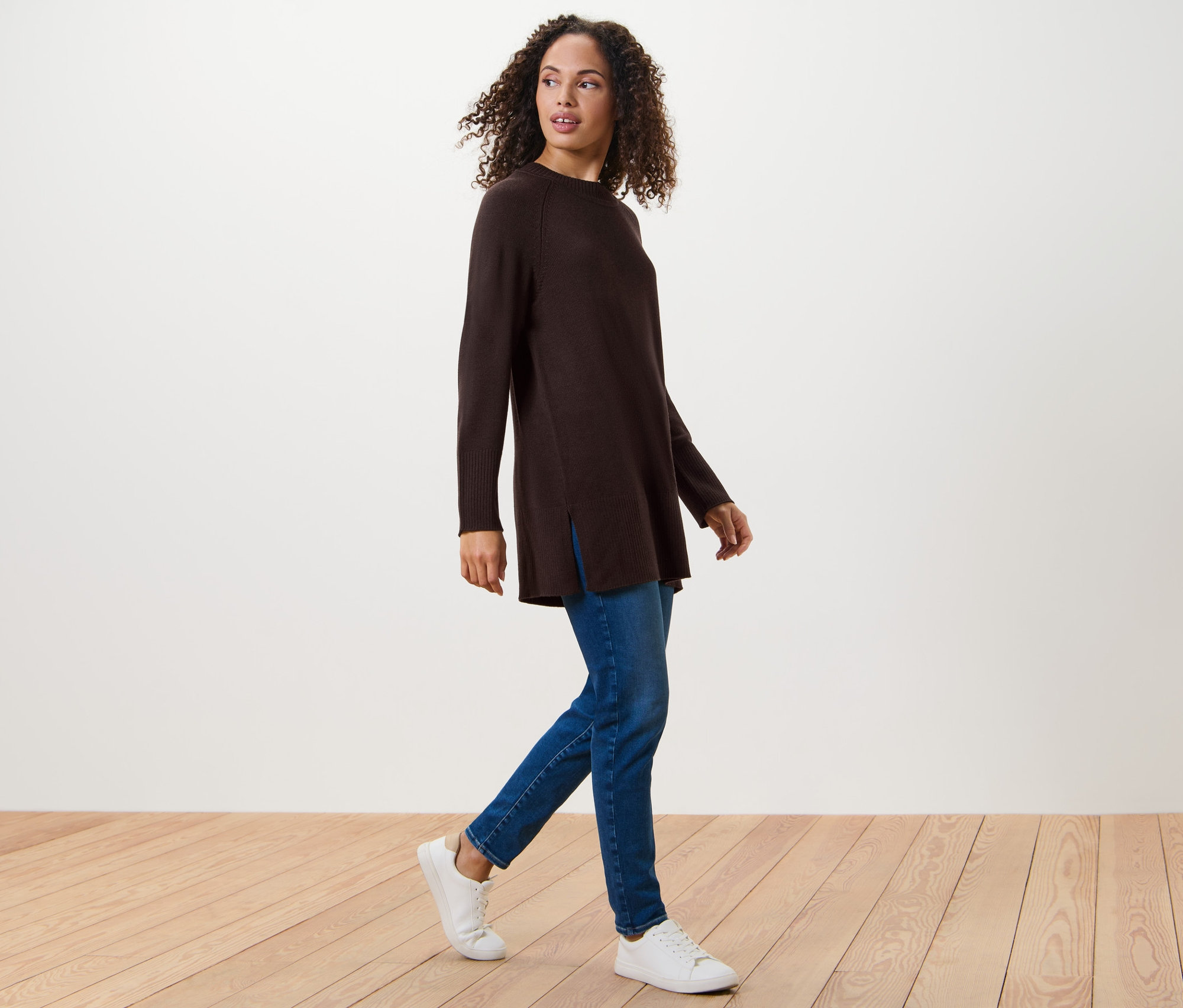 Žena kráčí a má na sobě dlouhý pletený svetr tmavě hnědé barvy, džíny a tenisky.
