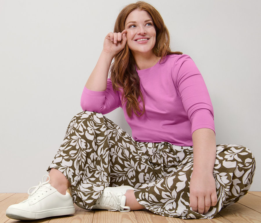 Žena sedí na podlaze v růžovém tričku s tříčtvrtečním rukávem a vzorovaných kalhotách.