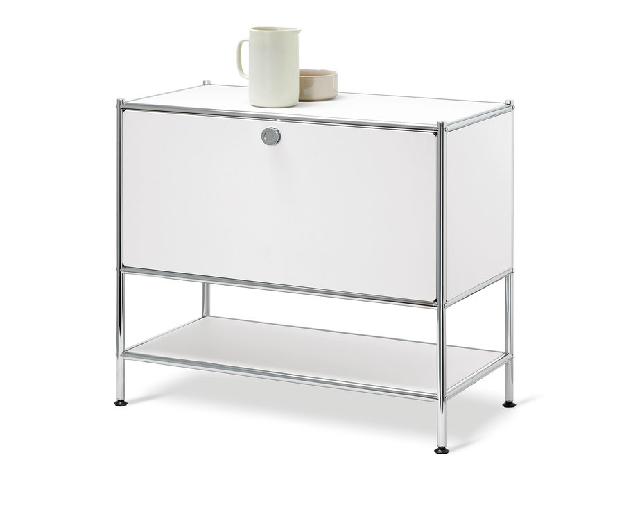 Bílá odkládací skříňka CN3 s jedněmi výklopnými dvířky. Na horní desce stojí keramický džbán a miska.