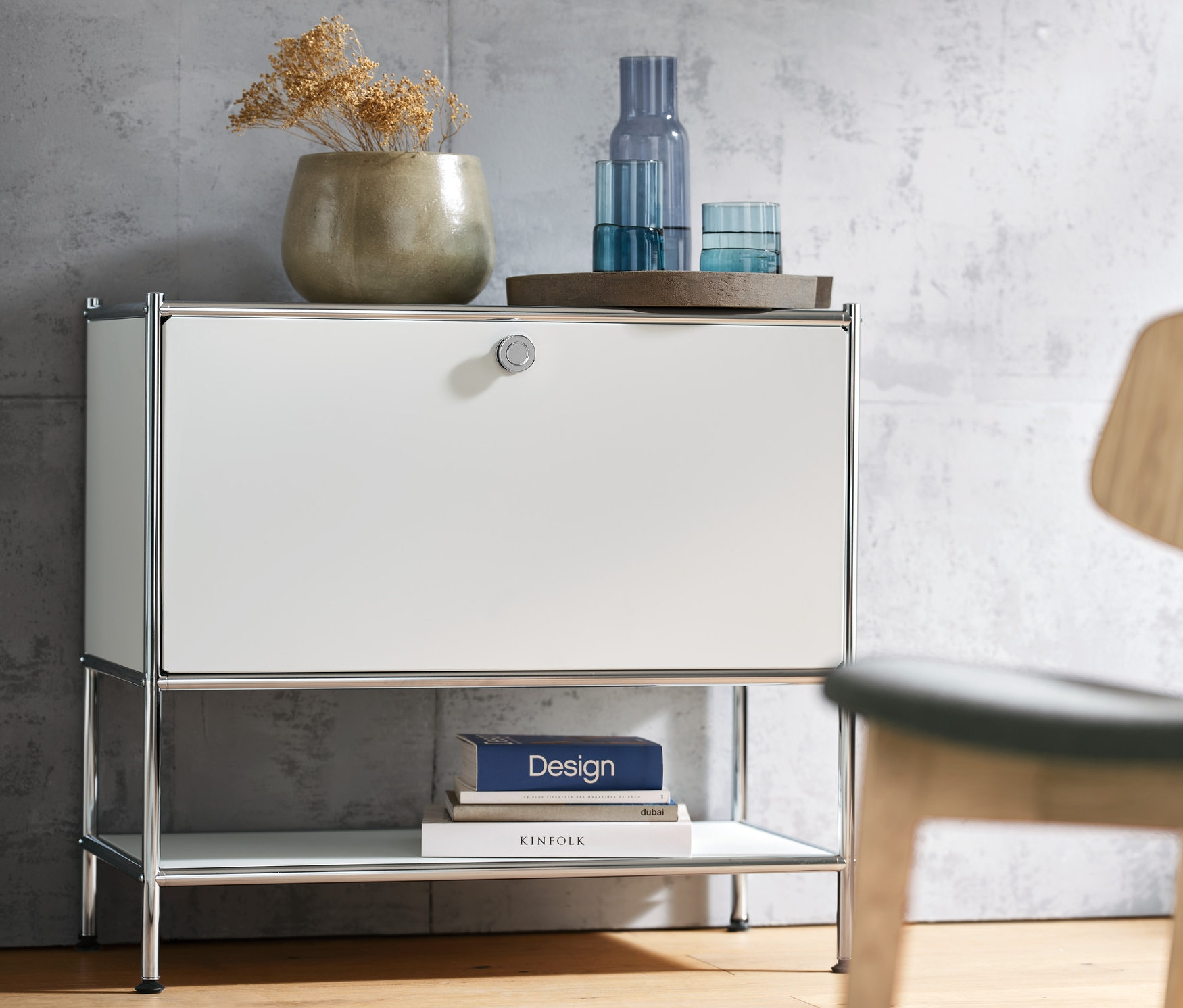 Bílá odkládací skříňka CN3 s jedněmi výklopnými dvířky, na ní váza a sklenice. Vpravo židle Lounge.