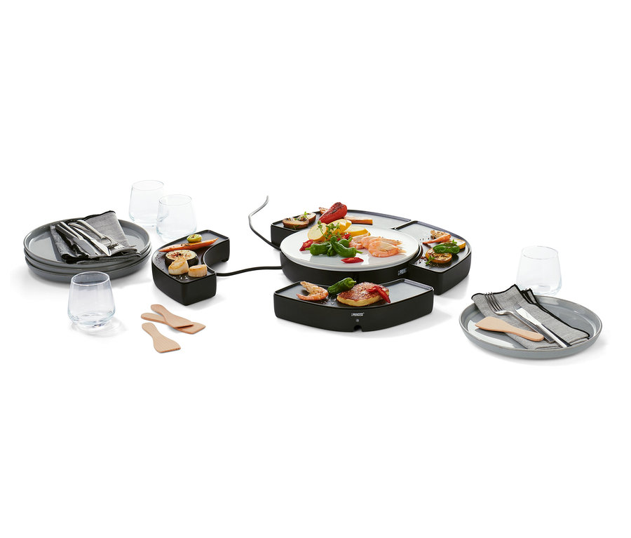Černý stolní gril »Dinner4All« s jídlem na grilovací ploše a talíři. Kolem jsou talíře s příbory, sklenice a dřevěné špachtle.
